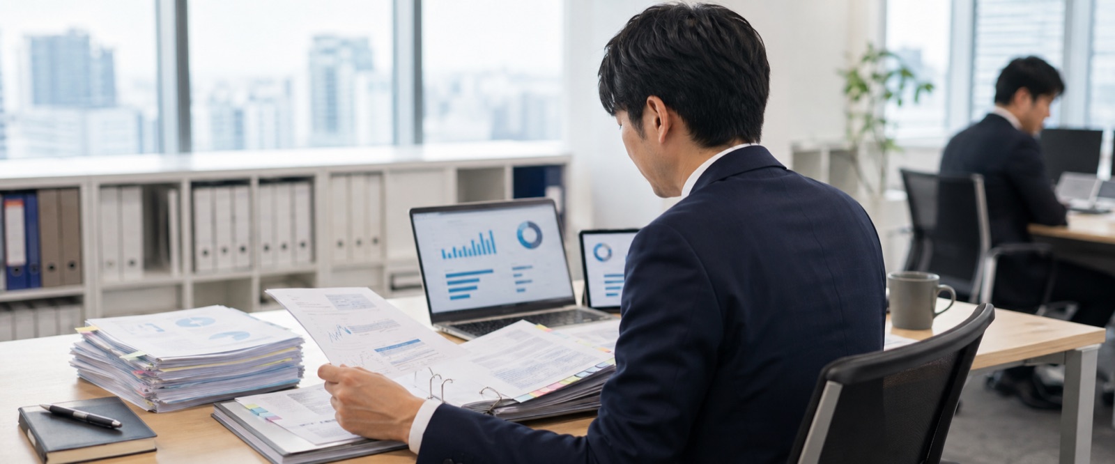 本部のデスクで、紙の日報、バインダー、ノートPCの一覧、タブレットの連絡を見比べながら各拠点の状況を確認している様子
